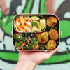 Hand holding a bento box with a variety of food items against a colorful background