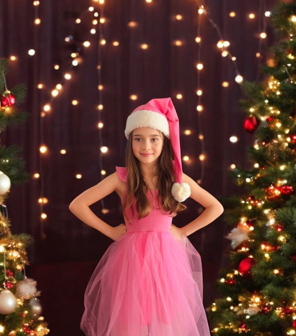 Young girl in a pink dress and Santa hat standing between two Christmas trees with lights in the background.