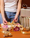 Person wrapping a gift with floral paper and gold ribbon on a table.