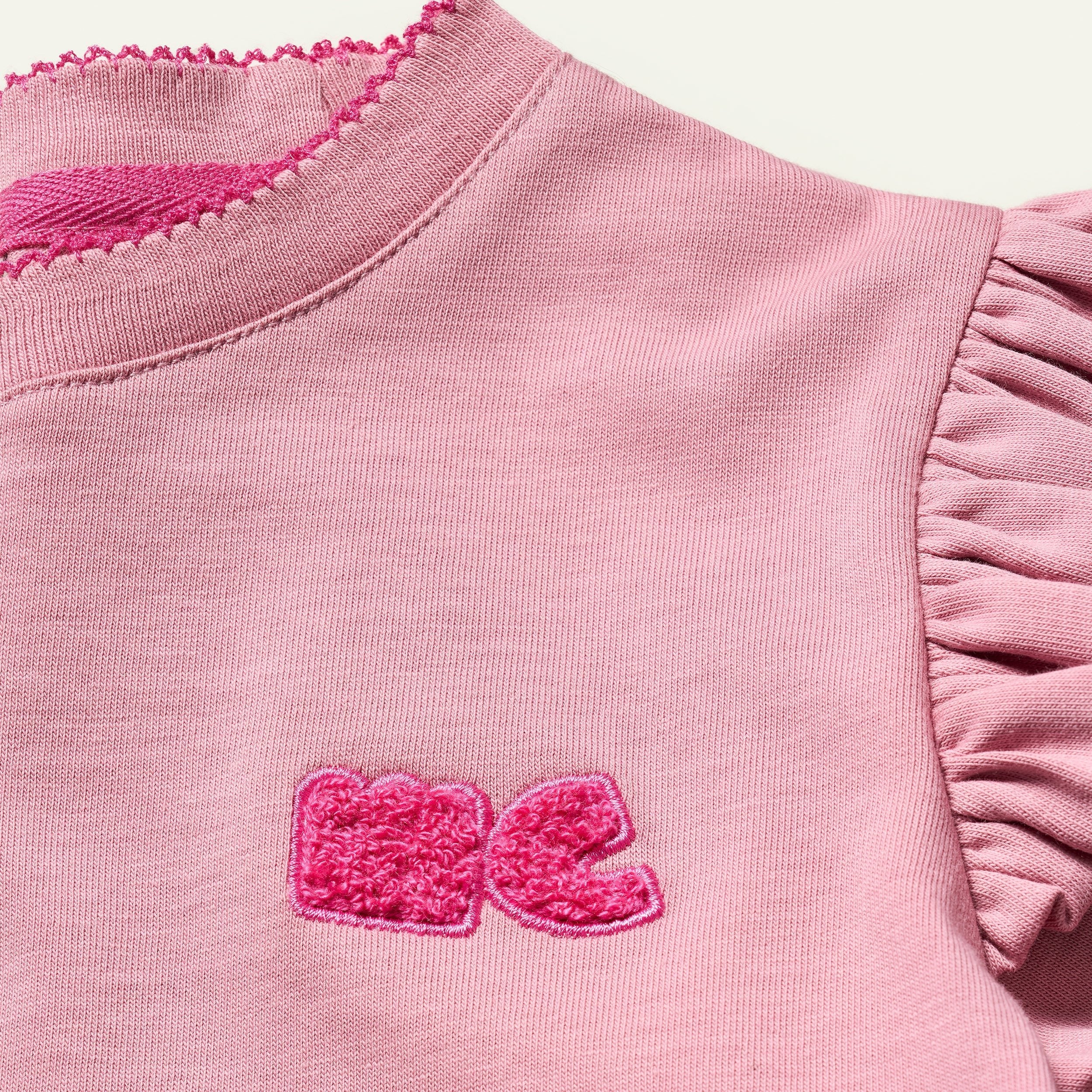 Close-up of a pink garment with ruffled sleeves and a logo on a light pink background