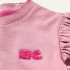 Close-up of a pink garment with ruffled sleeves and a logo on a light pink background
