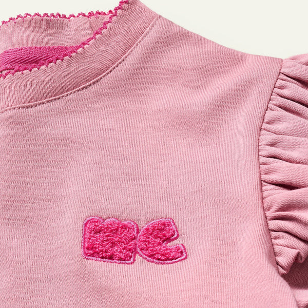 Close-up of a pink garment with ruffled sleeves and a logo on a light pink background