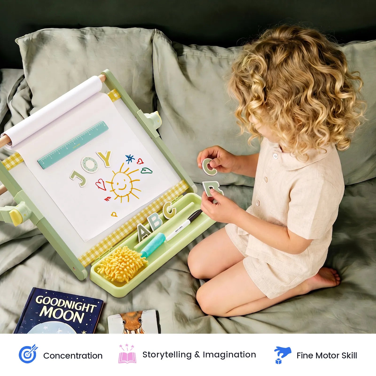 A young child with curly blonde hair sits on a gray couch next to a portable children's drawing board and tray set. The light green and yellow board has a white writing surface with colorful drawings, and the tray holds small toy pieces and accessories.