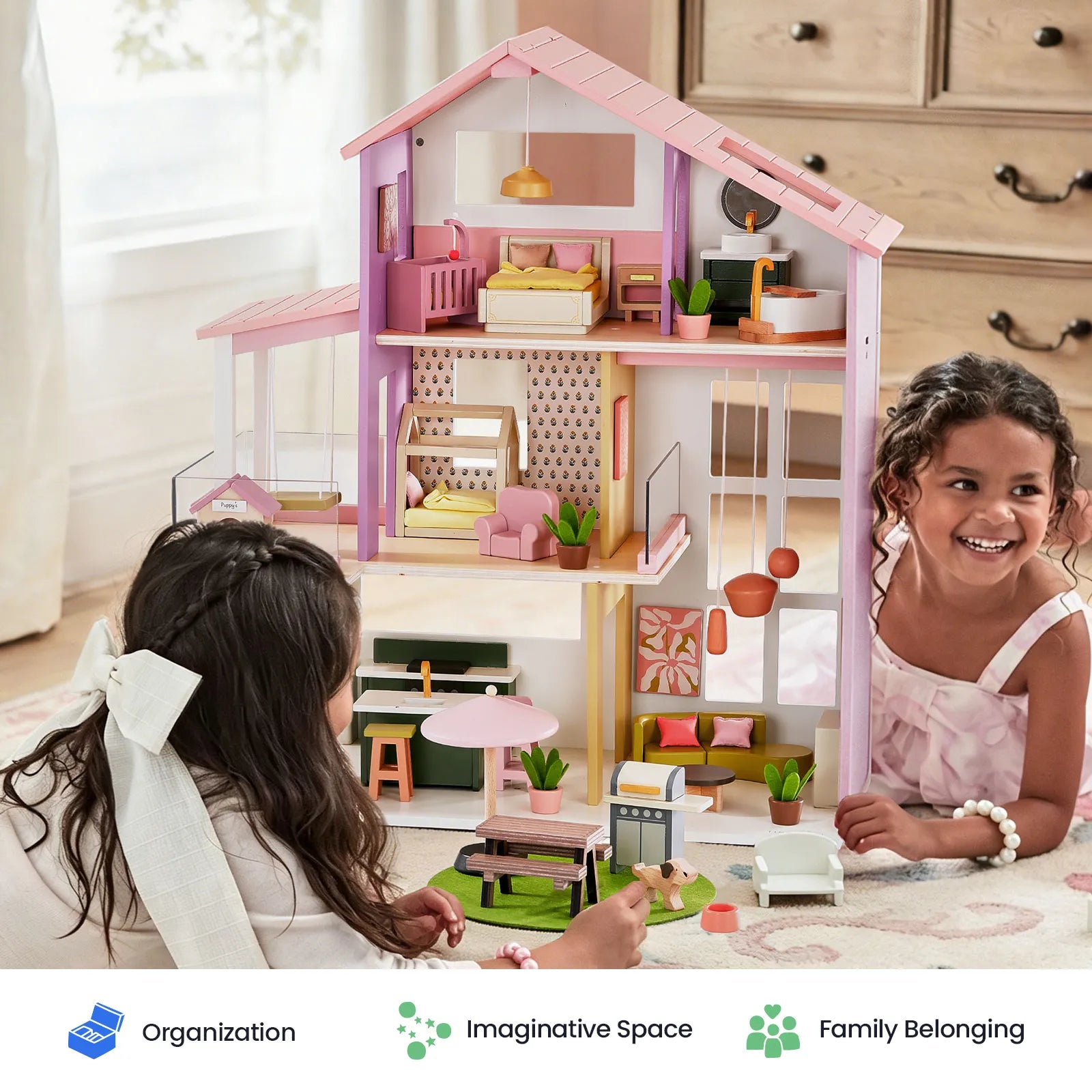 A large pink and purple wooden dollhouse with miniature furniture is shown in a bright room, with two children playing with it. Three text badges at the bottom read 'Organization', 'Imaginative Space', and 'Family Belonging'.