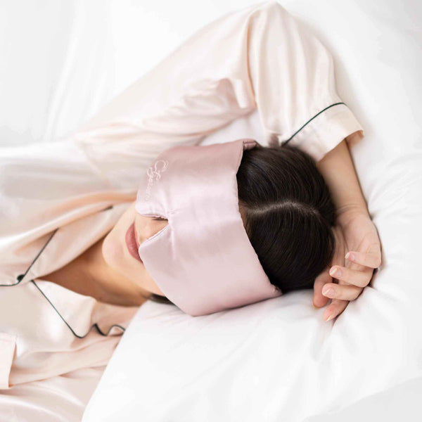 Person wearing a pink satin eye mask and pajamas, lying on a white bed.