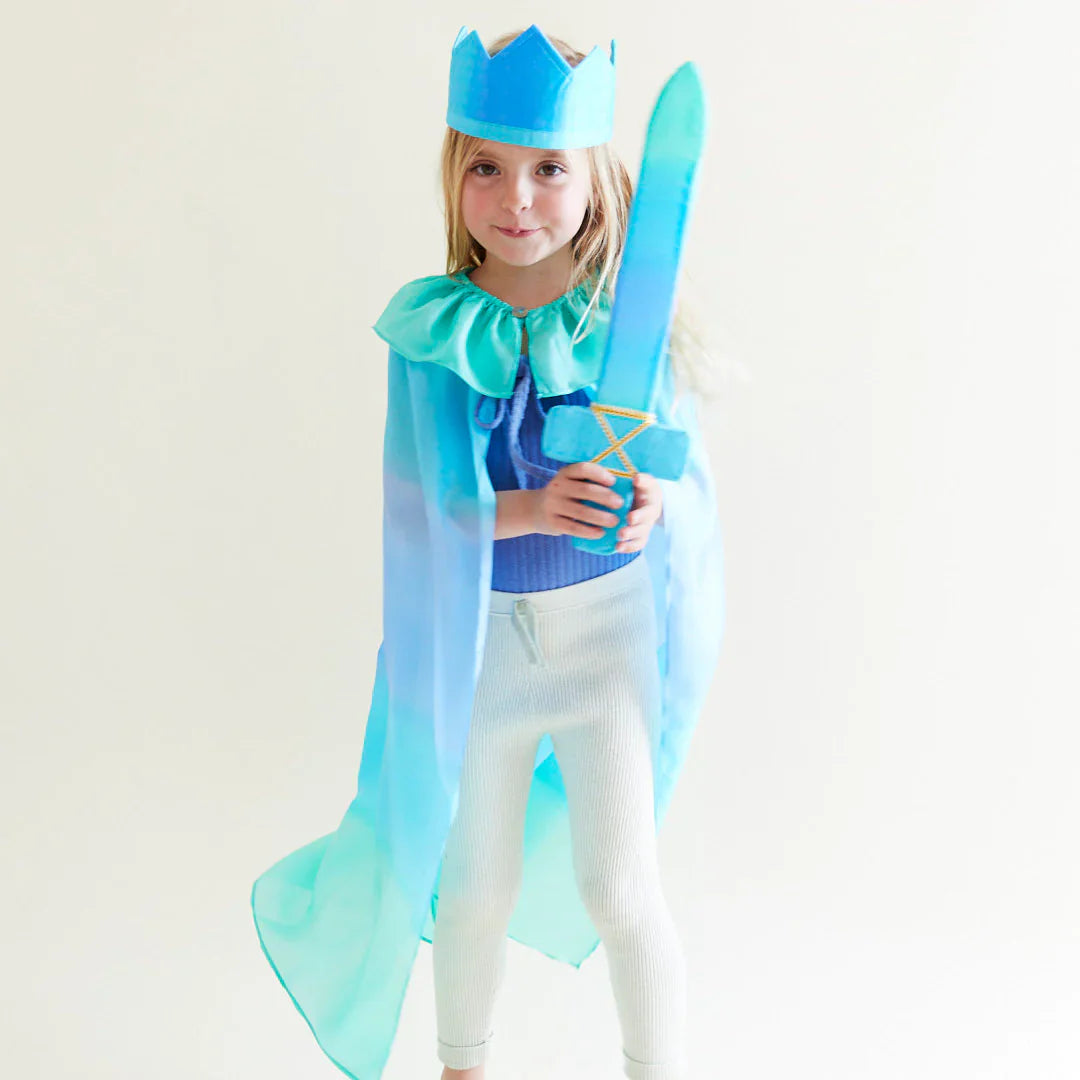 Child in a blue and green costume with a cape and crown, holding a sword on a white background