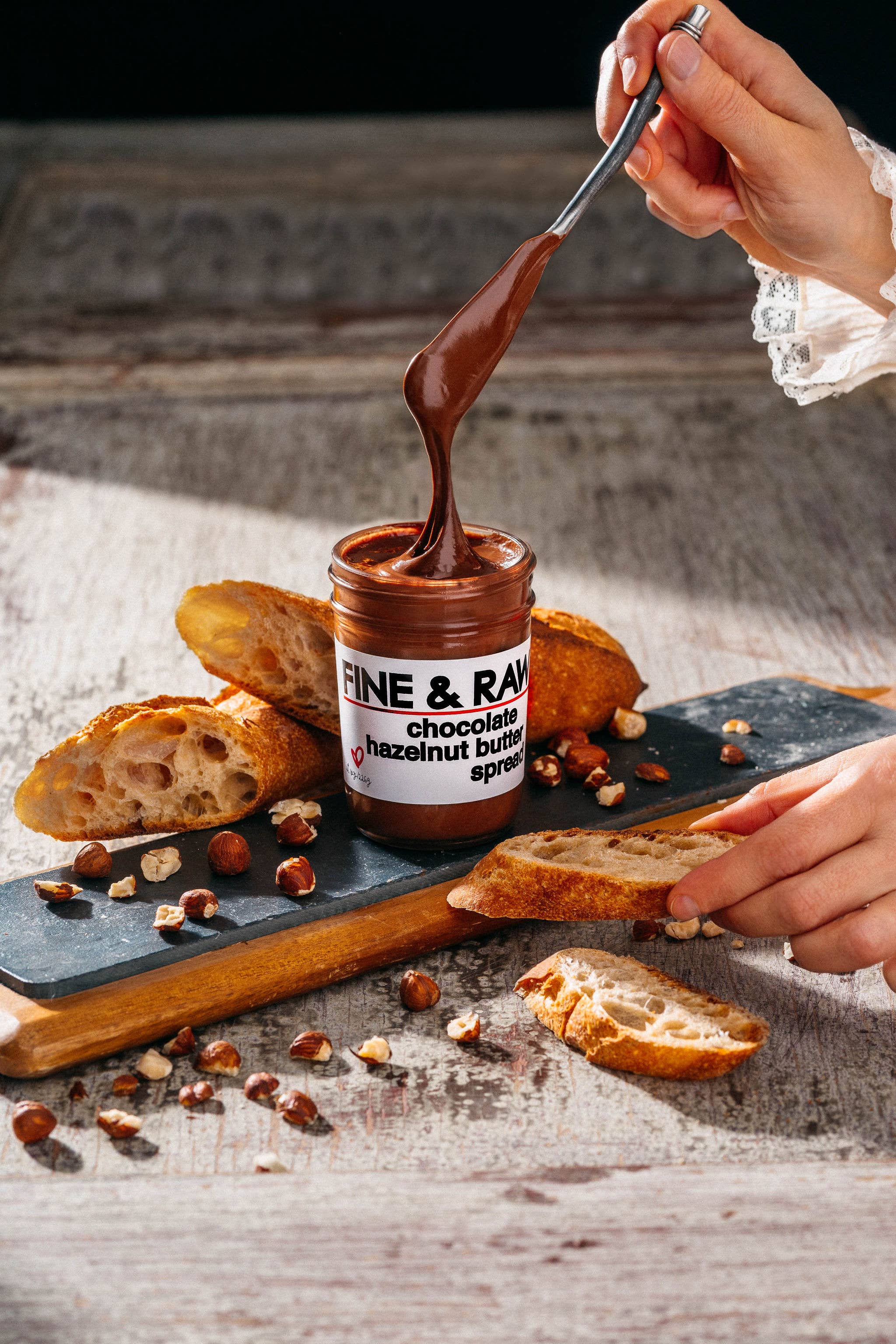 Jar of 'Fine & Raw' chocolate hazelnut spread being used to dip bread on a rustic wooden surface.