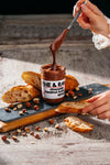 Jar of 'Fine & Raw' chocolate hazelnut spread being used to dip bread on a rustic wooden surface.