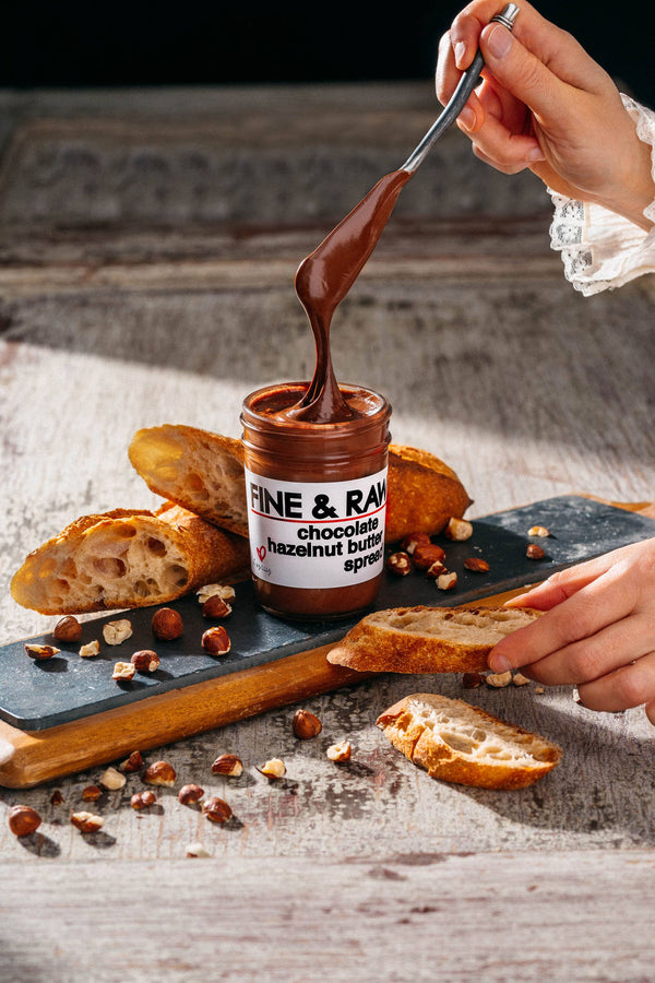 Jar of 'Fine & Raw' chocolate hazelnut spread being used to dip bread on a rustic wooden surface.