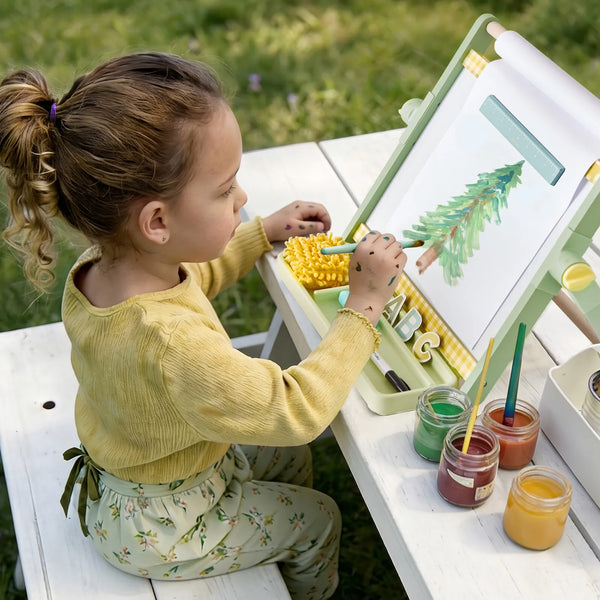 A young child sits on a white wooden bench outdoors, painting on a small easel. The scene includes paint jars, brushes, and a green palette on a white table in a grassy outdoor setting.