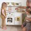 A toddler play kitchen set in a home setting, featuring a white and gold toy stove, oven, sink, and microwave with a patterned backsplash. A young girl in a brown patterned pajama set stands next to it, while a man and a baby watch from the right side of the frame.