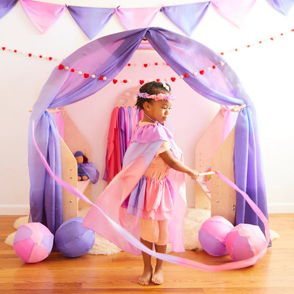 Child in a pink and purple dress with a cape standing inside a homemade play tent.