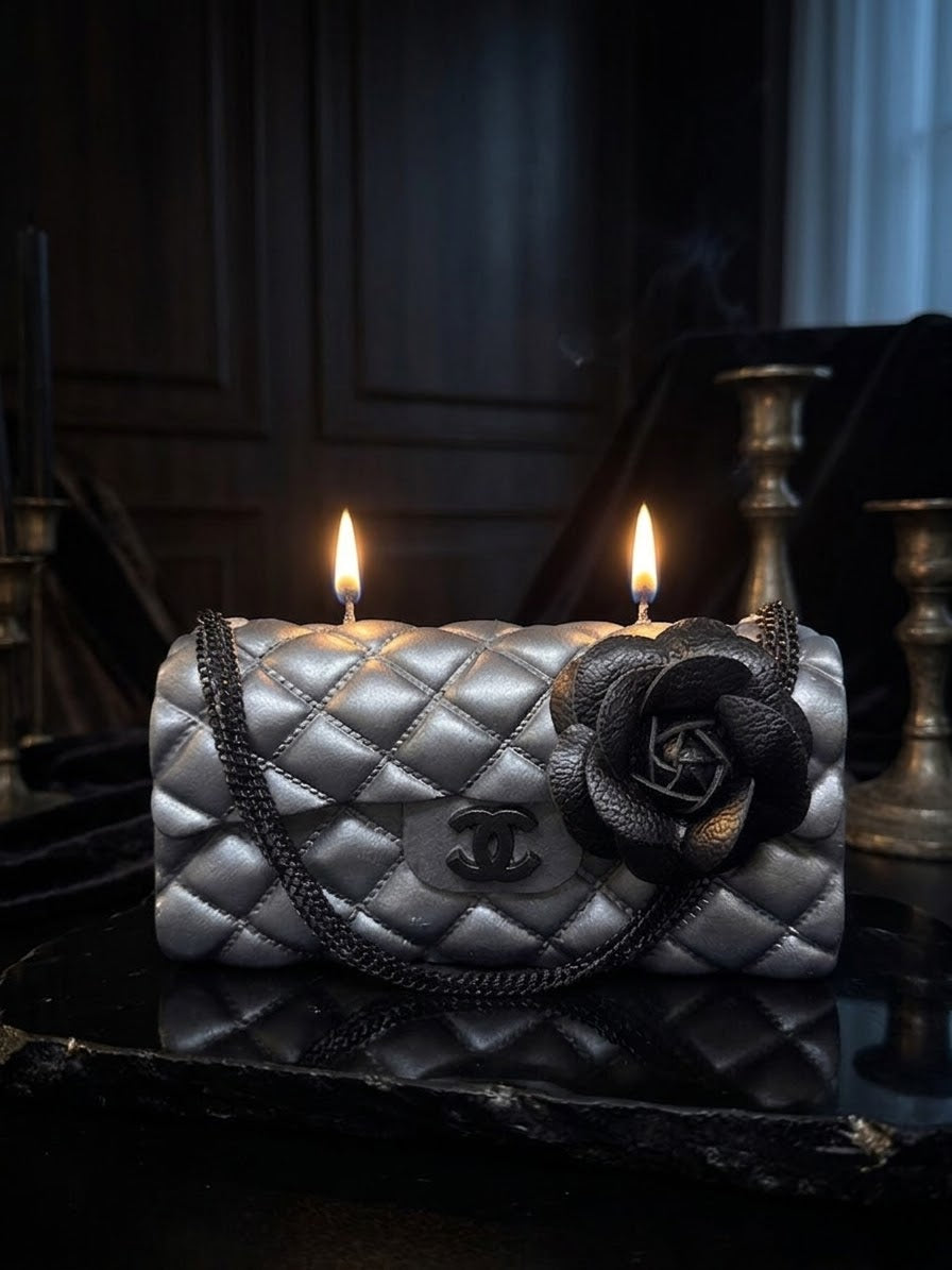 A double-wick candle styled as a quilted silver Chanel flap bag with a black leather rose accent and double flames, set on a dark reflective surface.