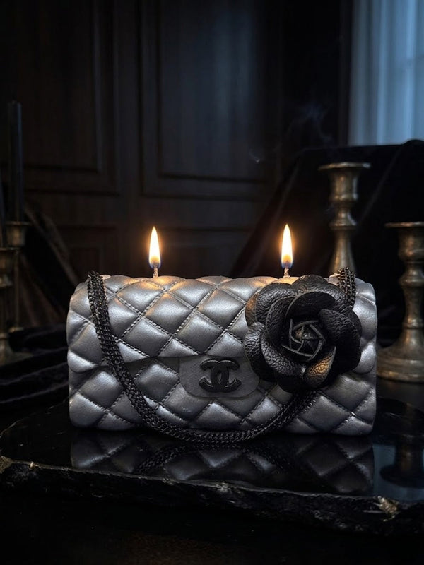 A double-wick candle styled as a quilted silver Chanel flap bag with a black leather rose accent and double flames, set on a dark reflective surface.