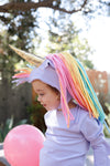 A toddler wearing a pastel lavender unicorn costume with a gold horn and long rainbow-colored faux hair, photographed outdoors with a pink balloon in the foreground.