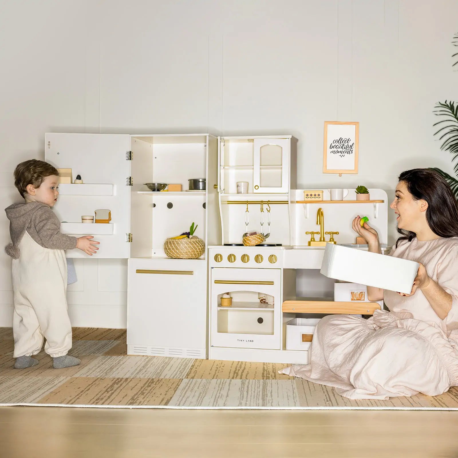 Children playing with a toy kitchen set in a room with a plant and framed picture on the wall.