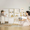 Children playing with a toy kitchen set in a room with a plant and framed picture on the wall.