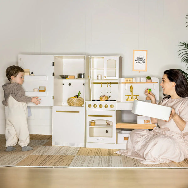 Children playing with a toy kitchen set in a room with a plant and framed picture on the wall.