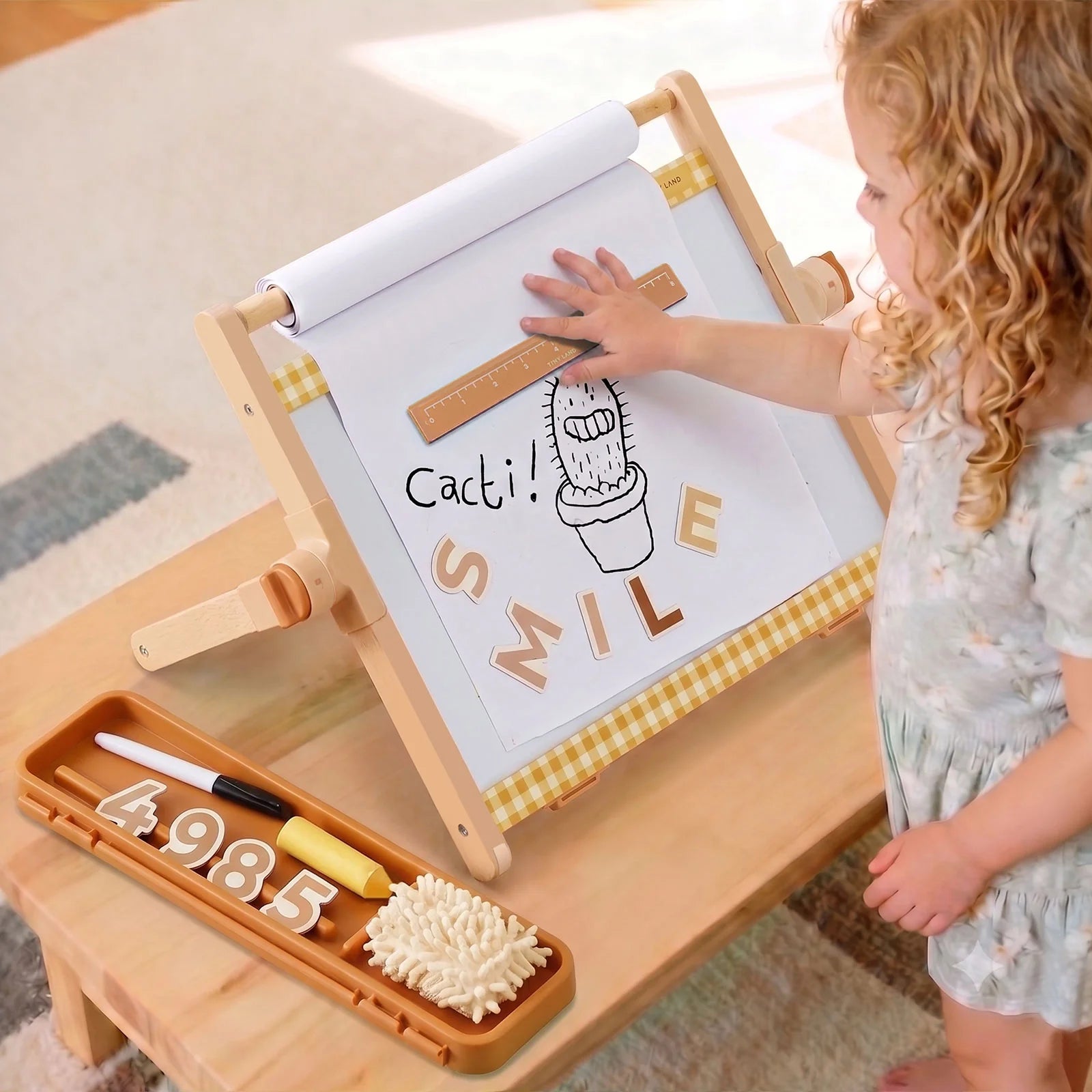 A young child stands beside a wooden activity board and tray, touching the board with a wooden stick. The board has a white writing surface with a cactus drawing and the words “Cactus Smile,” and the tray holds number tiles and a brush in a bright indoor setting.