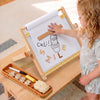 A young child stands beside a wooden activity board and tray, touching the board with a wooden stick. The board has a white writing surface with a cactus drawing and the words “Cactus Smile,” and the tray holds number tiles and a brush in a bright indoor setting.