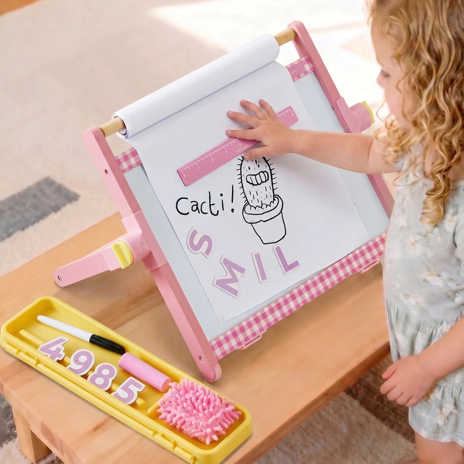 A young child stands beside a pink children's drawing easel on a wooden table, touching the white drawing surface. The easel has a pink-and-white gingham frame, a white roll of paper, and a yellow tray below holding paintbrushes, numbers, and a pink brush.