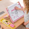 A young child stands beside a pink children's drawing easel on a wooden table, touching the white drawing surface. The easel has a pink-and-white gingham frame, a white roll of paper, and a yellow tray below holding paintbrushes, numbers, and a pink brush.