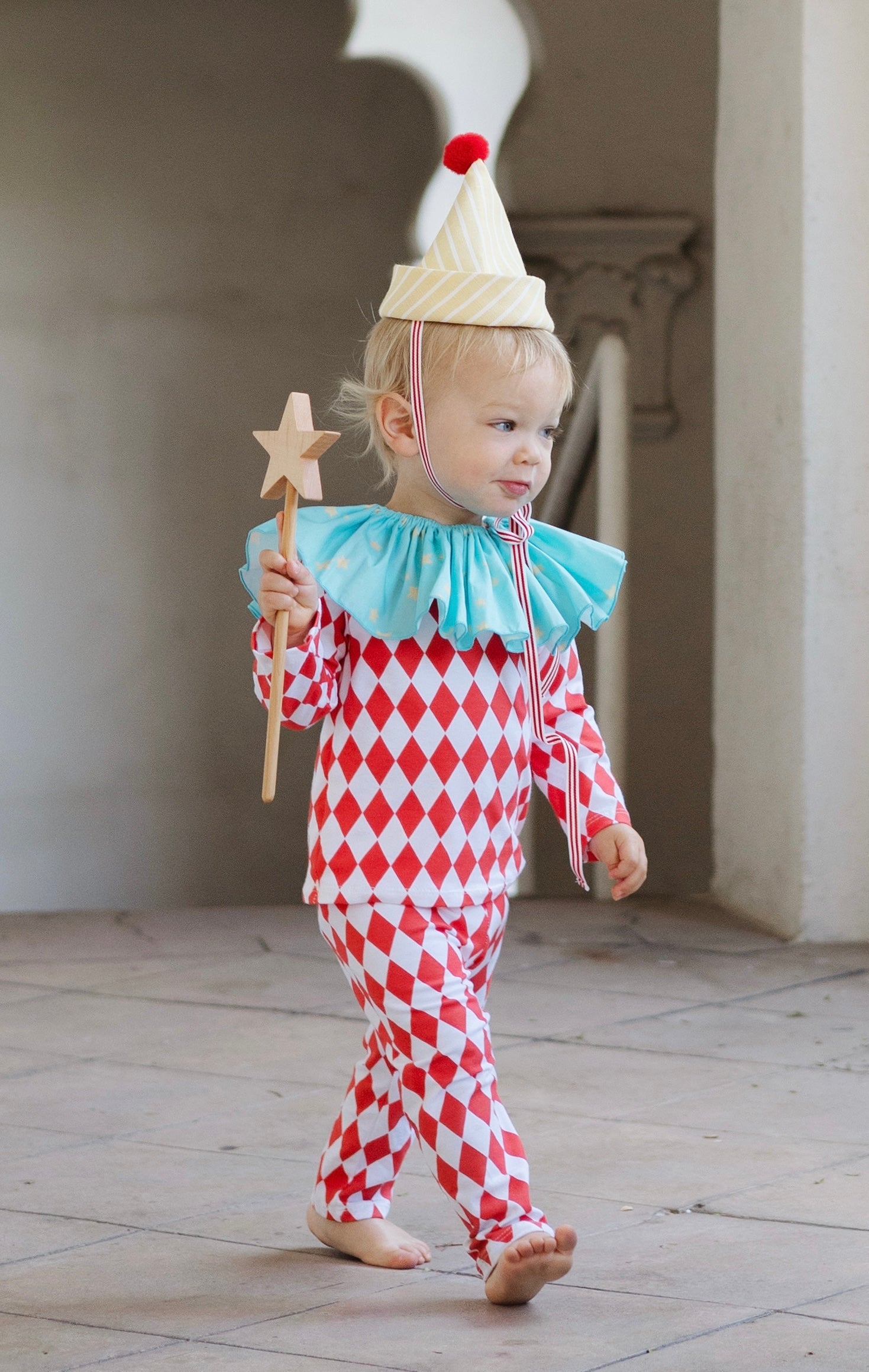 Child in a clown costume with red and white checkered outfit, blue ruffled collar, and white hat with red pom-pom.