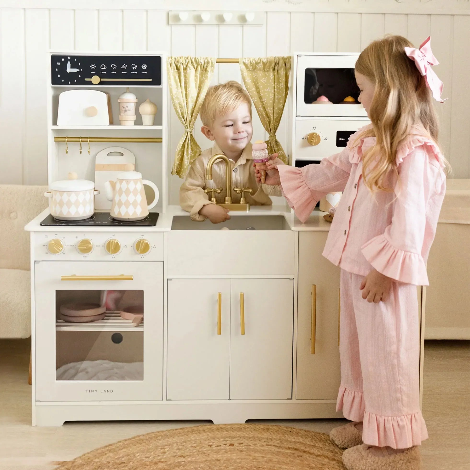 A toddler boy and a young girl play with a small white wooden toy kitchen set in a bright room. The kitchen includes gold-tone knobs, a sink, pretend stove, shelves with toy items, curtains, and a small toy oven.