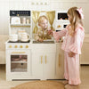 A toddler boy and a young girl play with a small white wooden toy kitchen set in a bright room. The kitchen includes gold-tone knobs, a sink, pretend stove, shelves with toy items, curtains, and a small toy oven.