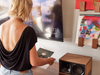 Woman interacting with a smart display device on a wall, with a wooden speaker in the foreground.