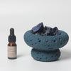 Blue volcanic ash bowl with dark stones and a bottle of essential oil on a white background