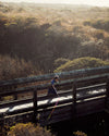 Person walking on a wooden boardwalk over a coastal landscape with trees and water.