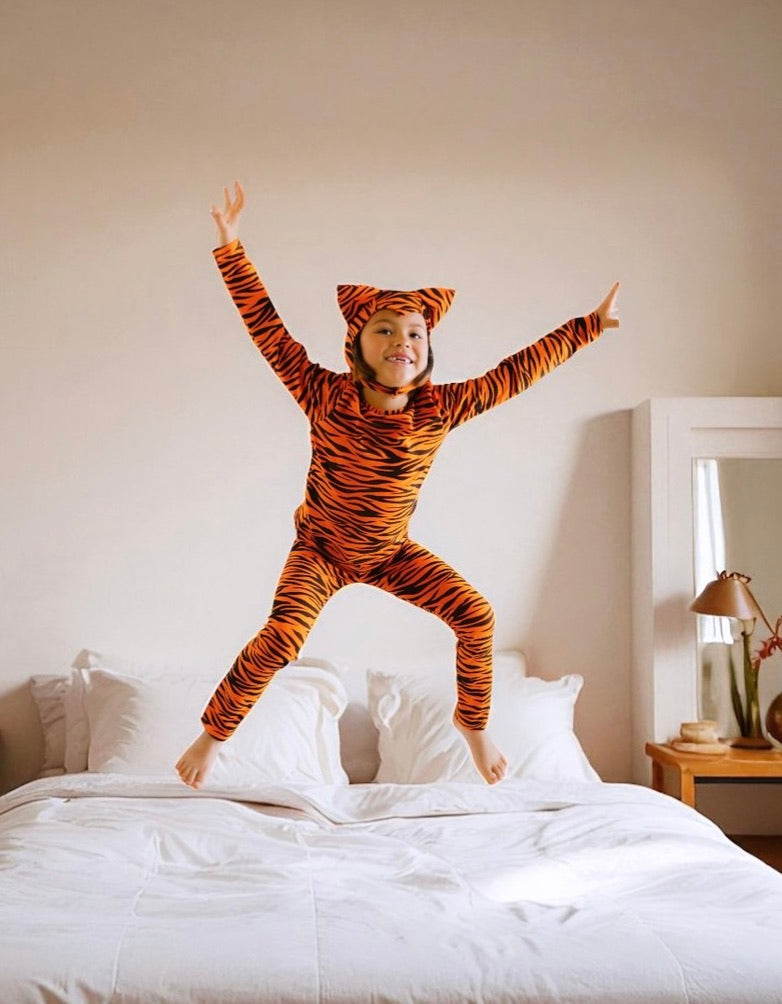 Child in a tiger-striped onesie jumping on a bed in a bedroom.