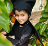 Child in a black bat costume among green leaves and fruits