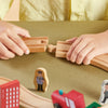 Two hands connecting wooden train tracks with small figures on a green surface.