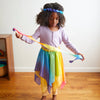 Child wearing a colorful rainbow skirt and headband, holding a ribbon, on a wooden floor.