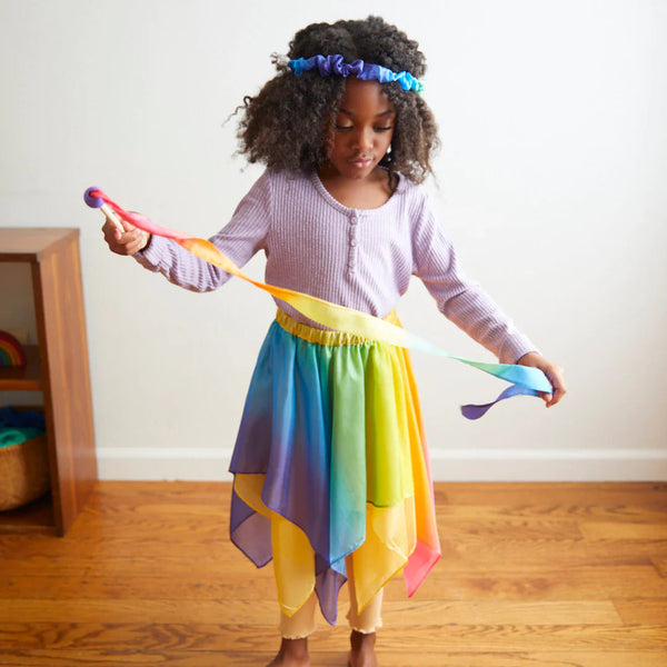 Child wearing a colorful rainbow skirt and headband, holding a ribbon, on a wooden floor.