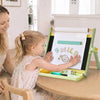 A young girl stands at a wooden table using a light green children's magnetic drawing easel, with a woman smiling beside her in a bright indoor room.