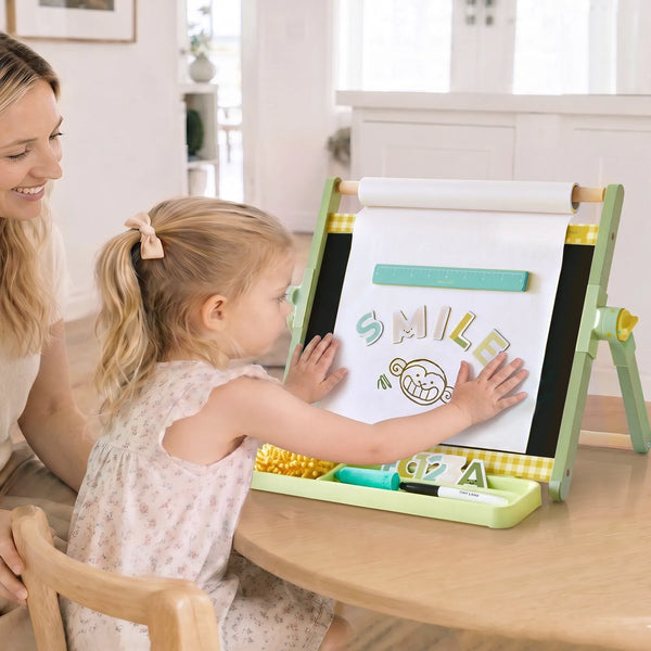 A young girl stands at a wooden table using a light green children's magnetic drawing easel, with a woman smiling beside her in a bright indoor room.