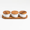 Three white jars with wooden lids on a wooden tray against a white background