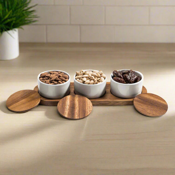 Three white bowls with snacks on a wooden tray on a kitchen counter.