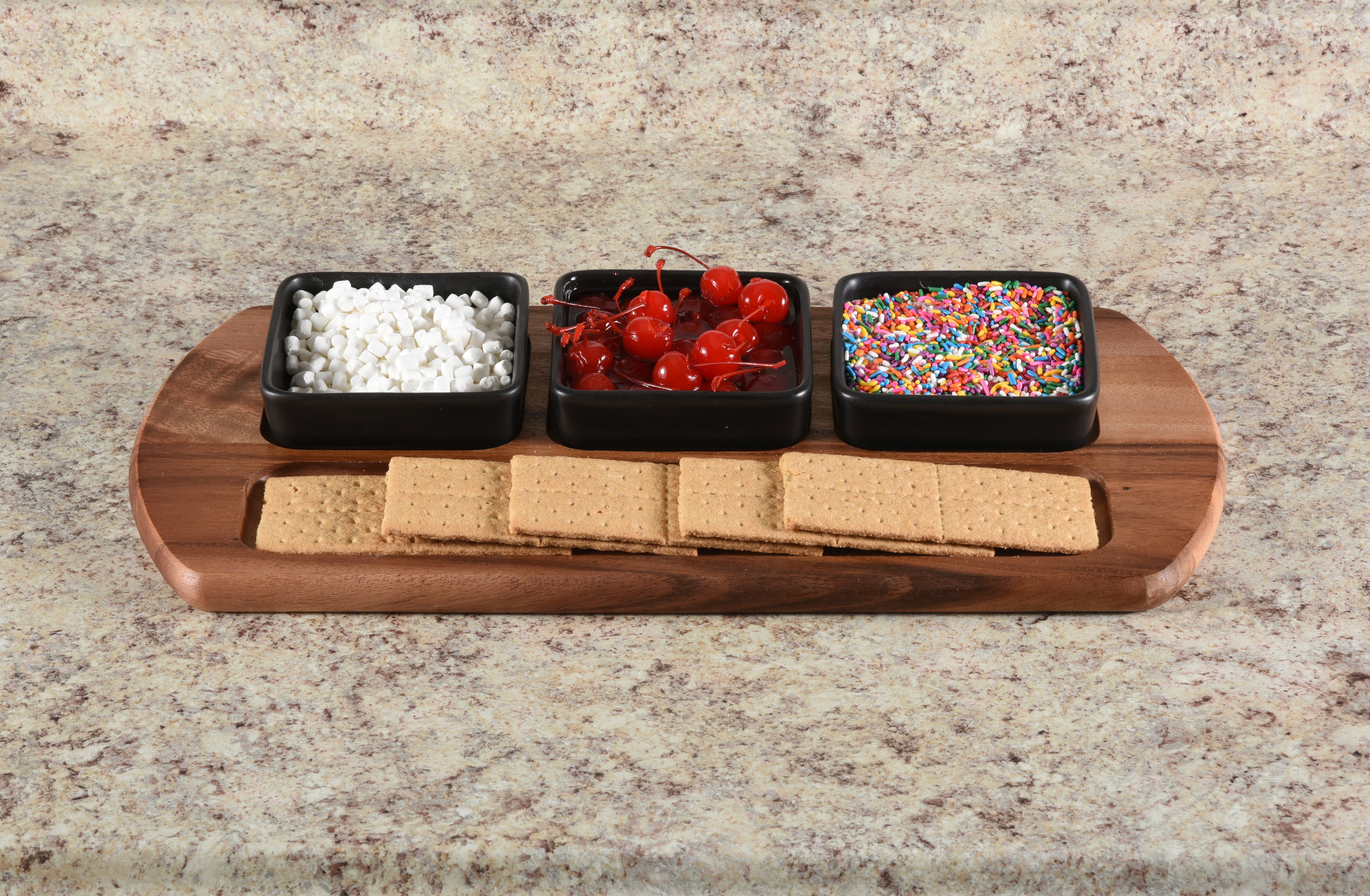 Wooden tray with graham crackers, white chocolate chips, red candies, and colorful sprinkles on a granite countertop.