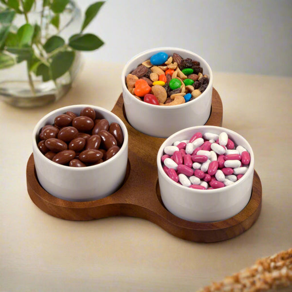 Three white bowls on a wooden tray with snacks on a light surface.