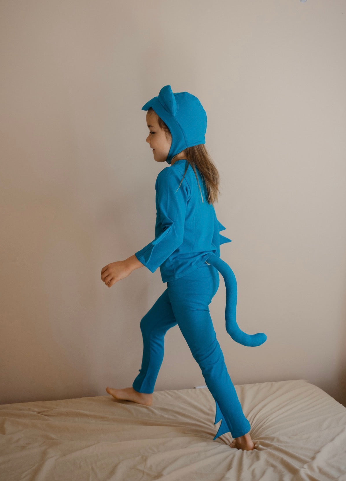 A child wearing a bright blue dinosaur costume, including a hood with ear-like flaps, a long-sleeve top, matching pants, and a tail, standing barefoot on a bed against a plain beige wall.