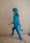 A child wearing a bright blue dinosaur costume, including a hood with ear-like flaps, a long-sleeve top, matching pants, and a tail, standing barefoot on a bed against a plain beige wall.