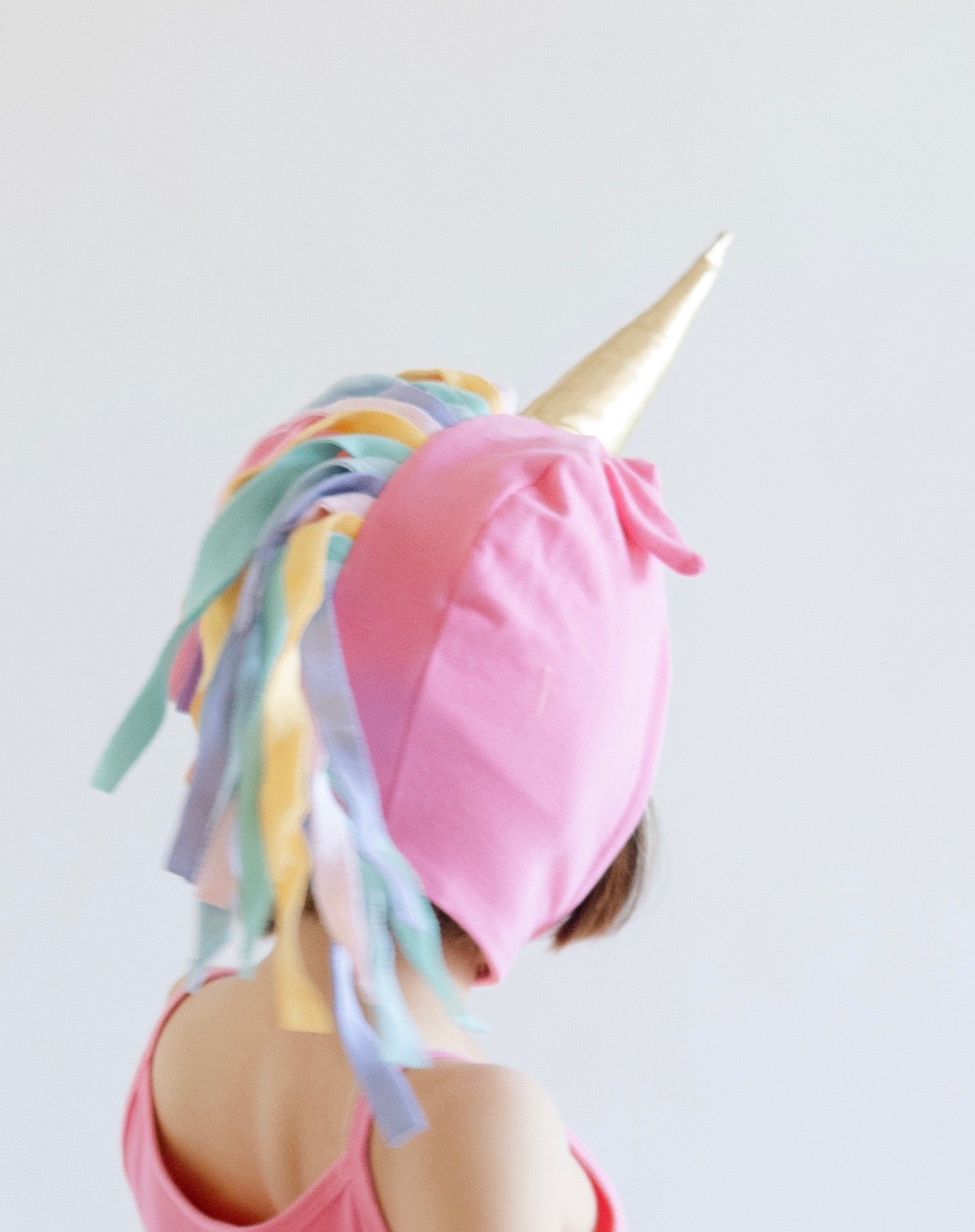 Person wearing a pink unicorn hat with colorful hair and gold horn on a light gray background