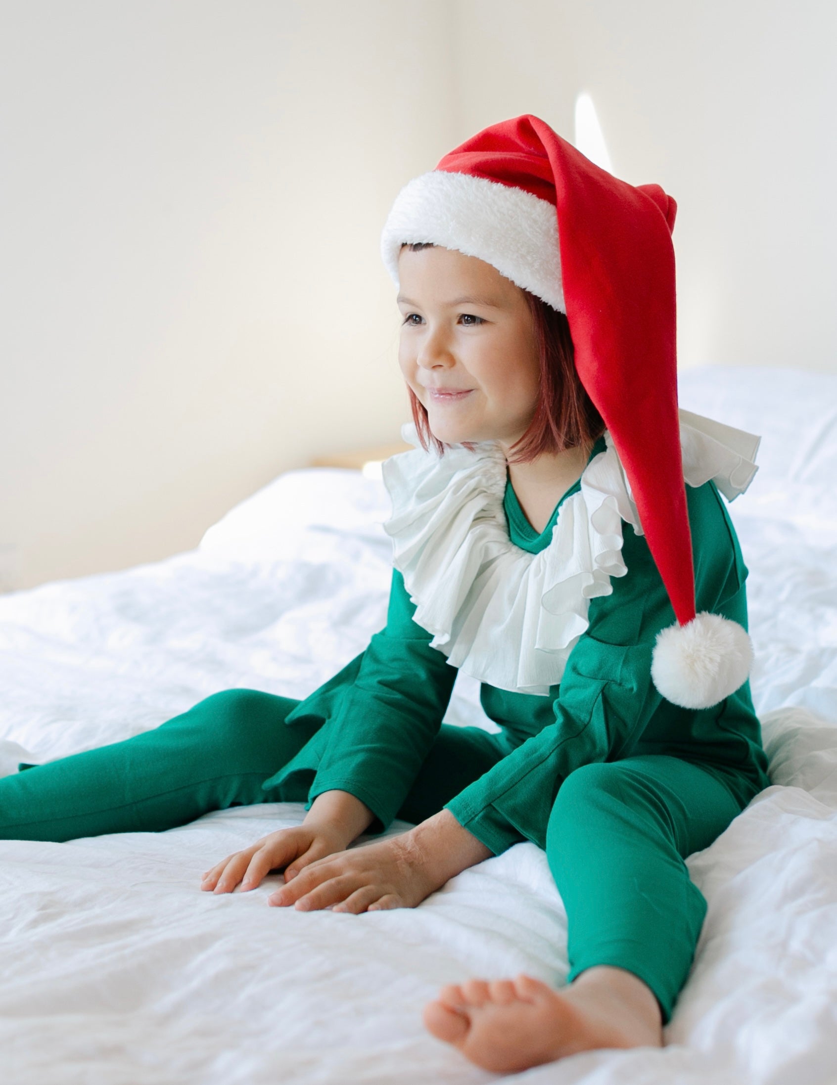 Child wearing a Santa hat and green elf outfit on a bed.