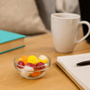 Glass bowl of colorful candies on a wooden table with a white mug, notebook, and pen in the background.
