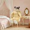 Stuffed toy in a yellow outfit standing in a bedroom with pink walls and white furniture.