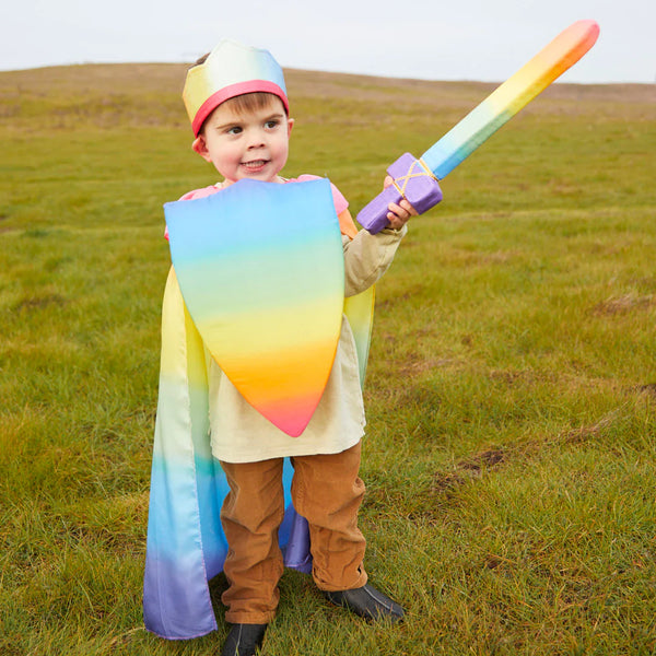 Child in a rainbow-themed costume holding a toy sword and shield in an open field.